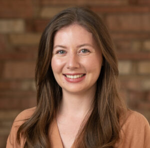 Kate McCabe, a person with brown straight hair, wavy over the shoulders, smiling at the camera with a brick background