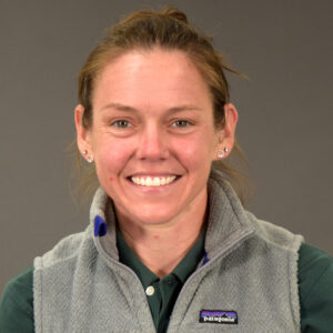 A person looks at the camera and smiles, wearing a gray vest with a gray background