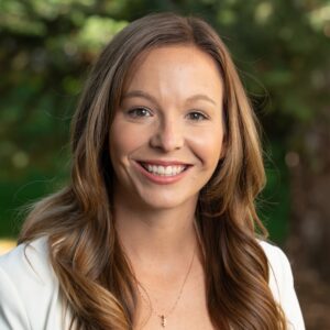 a woman smiles at the camera, with long brown hair and trees in the background