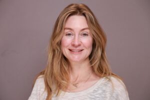 a person smiles at the camera, with blonde hair and a tan background