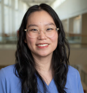 A person with long black hair and glasses smiles at the camera, the TMI in the background