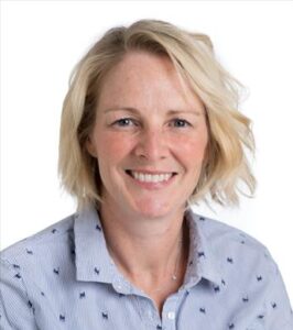 A person with short blond hair smiles at the camera, a white background and wearing a blue shirt