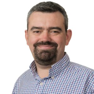 A person smiles at the camera, with brown hair and a brown goatee, a blue checkered shirt and a white background