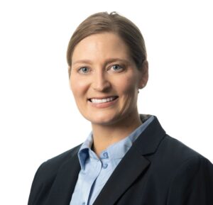 a person smiling at the camera with blond hair pulled back, a white background, and wearing a blazer jacket