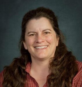 a person smiles at the camera with brown hair, a gray background, and an orange plaid shirt