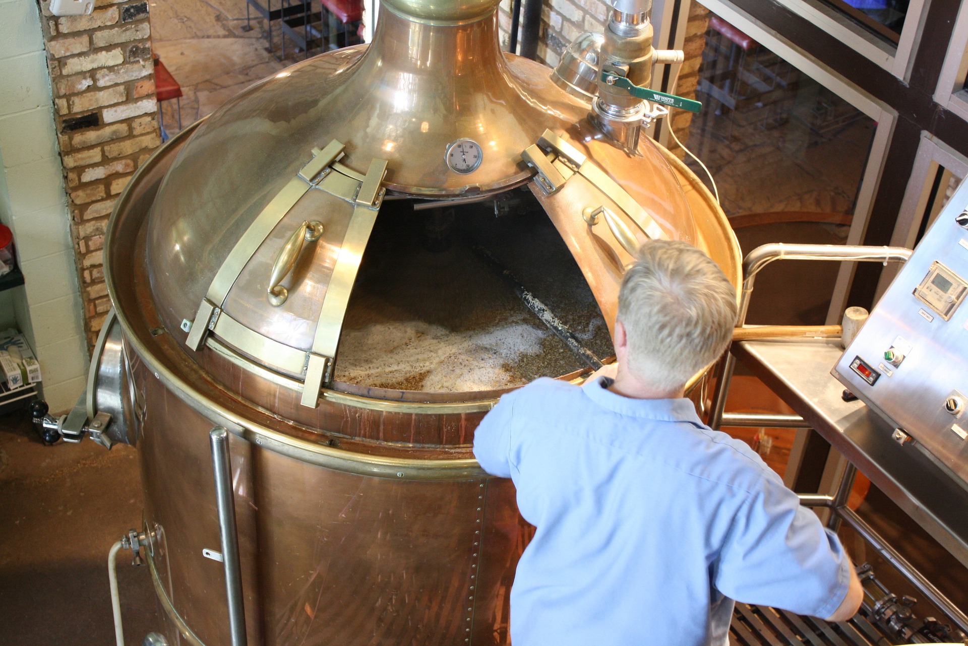 A large copper kettle. The top is open and a man in a light blue shirt uses a long handled tool to stir inside the kettle.