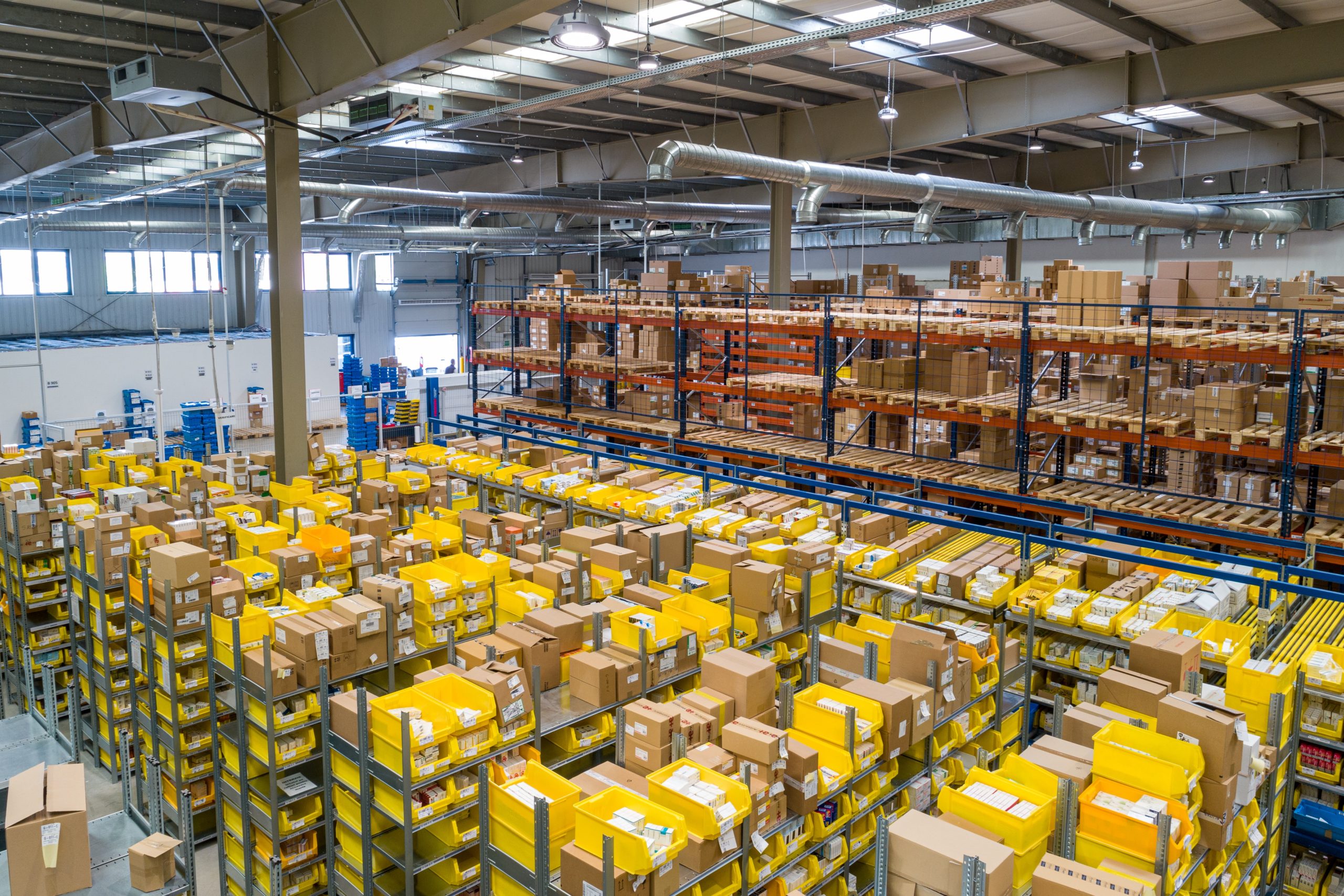 Birds eye view of a warehouse. In the foreground small cardboard boxes are in yellow contains. In the back, larger boxes are stacked on tall metal shelves.