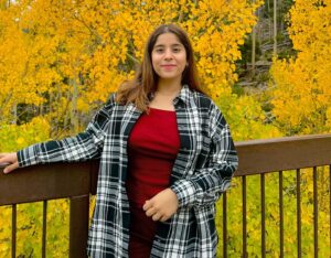 a person smiles at the camera on a porch, outdoors in the fall, with fall foliage in the background. She has long brown hair and is wearing a red shirt and a flannel over it.