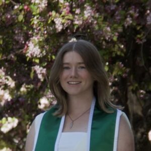 a person smiles at the camera with shoulder length brown hair and a graduation sash