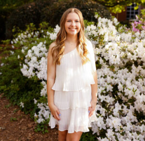 a person with long blond hair stands in front of a bush with white flowers, wearing a white dress and smiling at the camera