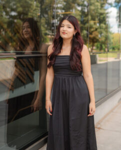 A person with long blackish-red hair smirks at the camera, wearing a long black dress and standing in front of windows reflecting the trees outside