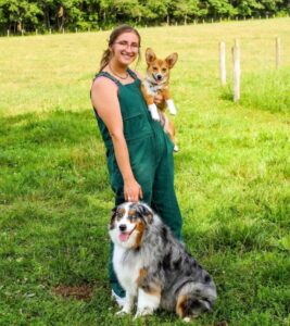 A person with long brown hair pulled back and dressed in green overalls, holds a tan corgi while holding on to a multi colored dog with a blue eye and a brown eye. Smiling at the camera in a green field.