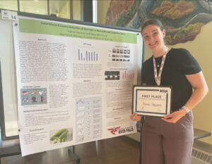 A person with brown hair smiles at the camera, standing in front of a board they made that one first place. They're holding a first place certificate.