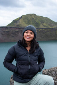 A person with black hair and a dark grey stocking cap, stands in front of a lake with a mountainous background, smiling, with her hands in the pockets of her black jacket.