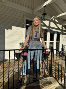 a person with long blond hair stands on a porch, smiling, with a college t-shirt on that has a purple "T"