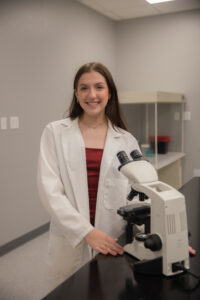 A person stands in a lab in a white coat, next to a microscope. The person has long brown hair, swooped behind her shoulders and is smiling at the camera.