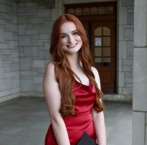 A person with long red hair smiles at the camera, wearing a red dress and clutching a graduation cap, a stone entry way in the background