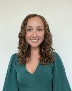 A person with green eyes and shoulder-length curly brown hair smiles at the camera, wearing a green shirt.