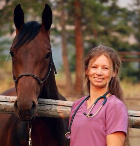 A person in pink scrubs stands next to a fence, she has long brown hair and is smiling at the camera. She is holding the reigns of a chestnut colored horse