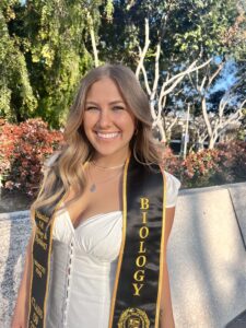 A person smiles at the camera, with wavy blond hair, wearing a white dress with a black and gold sash and trees in the background