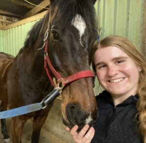 A person smiles at the camera, taking a selfie with a brown and white horse, the green stall in the background