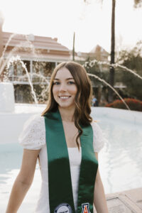 a person with brown hair stands in front of a fountain, smiling in a white dress, with a CSU sash over her shoulders