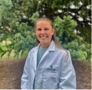 a person in a white doctor's coat smiles at the camera, outside with summer foliage in the background