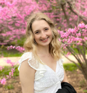 A person smiles at the camera, in a white dress, with blonde hair wavy around her face, a tree with pink blossoms in the background.