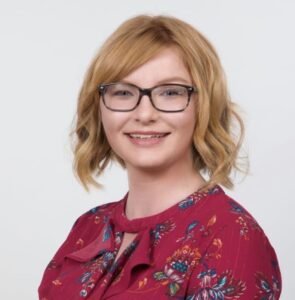 A person with short red hair framed around their face and black glasses, smiles at the camera wearing a red flowery shirt and plain white background