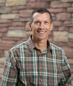 a person smiles at the camera, wearing a plaid shirt with a red and white brick wall in the background