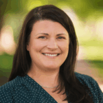 a person smiles at the camera with dark brown hair and a green summer background.